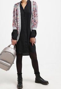 Schwarzes knielanges Kleid mit langen Ärmeln, kombiniert mit einer mehrfarbigen Strickjacke und schwarzen Schnür-Stiefeln. Silberner Rucksack.