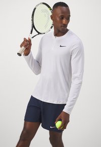 Ilga rankovė balta sportinė marškinė su užtrauktuku ir juodu Nike logotipu, derinama su tamsiai mėlynomis šortais. Laiko teniso raketę ir kamuoliuką.