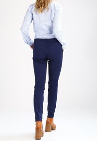 Navyblauwe fitted broeken met een gladde textuur en zakken, gecombineerd met een lichtblauw gestreept shirt. Draagt beige hakken.