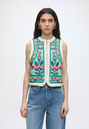 Donna che indossa un gilet smanicato color verde turchese con ricamo floreale colorato e jeans azzurri chiari, in piedi su uno sfondo bianco semplice.