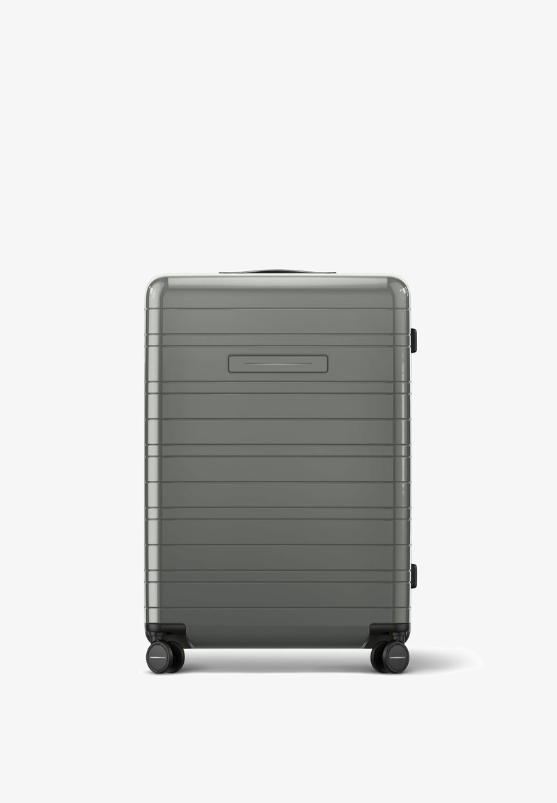 Valise rigide grise avec des rainures, une poignée supérieure et quatre roulettes. Elle dispose d'un petit espace pour une étiquette d'identification et d'une finition lisse et brillante.