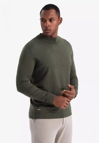 Maglione in maglia verde oliva con colletto alto, maniche lunghe e design aderente. Presenta una texture sottile e un logo vicino all'orlo.