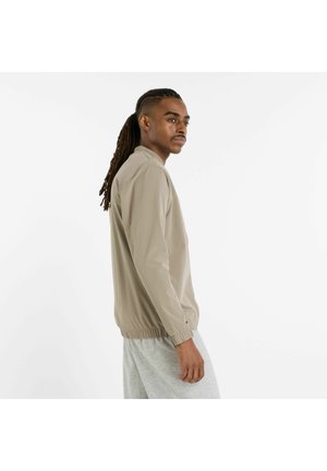 Beige letvægtsjakke med ståkrave, elastiske manchetter og kant, iført over grå shorts; glat tekstur og minimalistisk design.