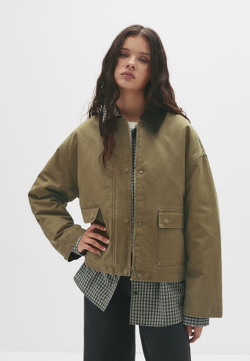 Khaki oversize jakna z gumbom, dvema sprednjima žepoma in korduisom ovratnikom, plastena čez karo srajco. Mat tekstura tkanine.