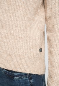 Beige gebreide trui met ribbelstructuur en zoom, met een klein zwart merketiket aan de zijkant. Gepaarde met donkerblauwe jeans.