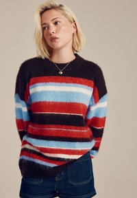 Striped sweater in shades of red, blue, and brown, with a soft, fluffy texture and a ribbed neckline. Paired with dark denim shorts.