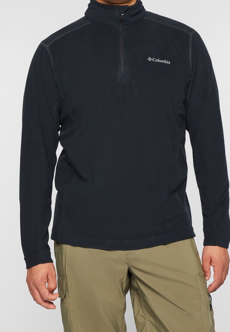 Mies, joka on pukeutunut mustaan Columbia vetoketjulliseen huppariin ja khakivärisiin cargo-housuihin, seisoo kädet rentoina sivuilla.