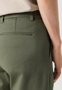 Gros plan d'une personne portant un pantalon vert olive avec une poche passepoilée à l'arrière, un haut beige rentré à l'intérieur, et son bras droit visible.