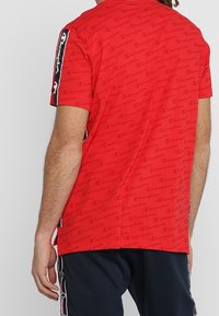 Rød kortærmet t-shirt med et heltrykt logo-mønster, sorte sidelommer og runde udskæring. Lavet af et blødt, åndbart materiale.