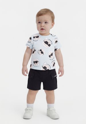 Copil mic cu păr castaniu deschis, purtând un tricou alb cu fețe de desene animate și pantaloni scurți negri cu buzunare, stând pe un fundal alb.