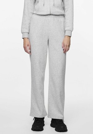 Dolna część ciała osoby noszącej jasnoszare spodnie dresowe z elastycznym pasem oraz czarne masywne sneakersy, stojącej na białym tle.