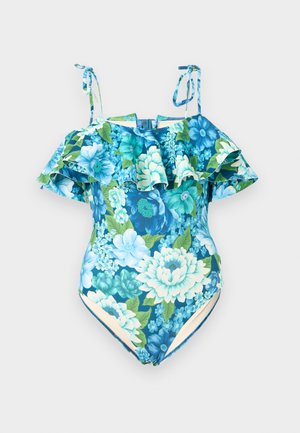 Mujer de pie descalza, con un bañador de una pieza azul con estampado floral, mangas con volantes fuera de los hombros y tirantes atados, fondo neutro.