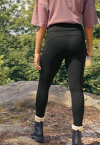 Mustad kõrge vöökohtadega mustad leggingid, millel on sile tekstuur ja tagataskud, kombineerituna suurte mustade saapadega, millel on valged kudumised ääred.