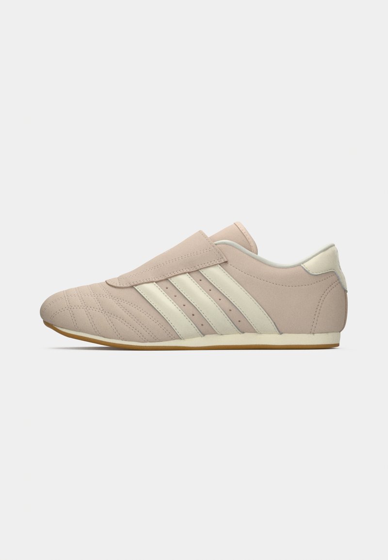 Zapatilla beige con franjas crema, talón acolchado, detalles de costura y suela de goma plana, mostrada de lado sobre un fondo blanco.