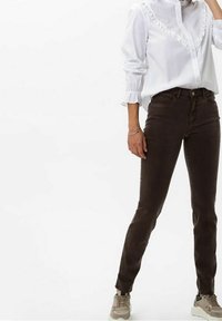 Žena v bílé blůze s dlouhým rukávem a volány, tmavě hnědých slim-fit kalhotách a béžových teniskách, stojící na bílém pozadí.