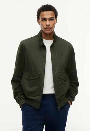 Junger Mann trägt eine dunkelgrüne Bomberjacke über einem weißen Hemd und dunkelblauen Hosen, steht mit den Händen in den Taschen vor einem einfarbigen Hintergrund.