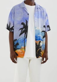 Persona de piel negra que lleva una camisa de botones de manga corta con un diseño de palmeras y un atardecer en la playa, una camiseta blanca y pantalones blancos.