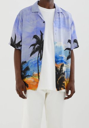 Persona de piel negra que lleva una camisa de botones de manga corta con un diseño de palmeras y un atardecer en la playa, una camiseta blanca y pantalones blancos.