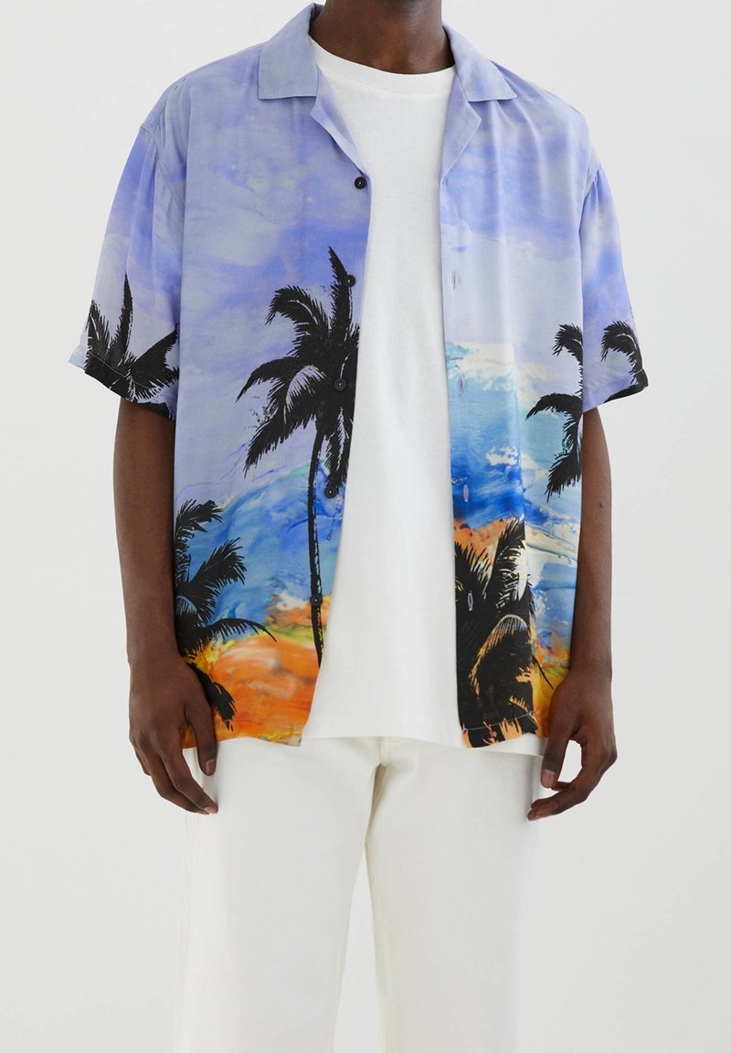 Persona de piel negra que lleva una camisa de botones de manga corta con un diseño de palmeras y un atardecer en la playa, una camiseta blanca y pantalones blancos.