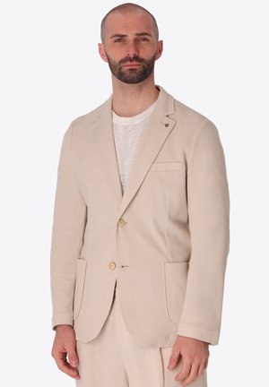 Uomo con barba corta che indossa un blazer beige chiaro strutturato sopra una t-shirt bianca, in piedi davanti a uno sfondo uniforme.