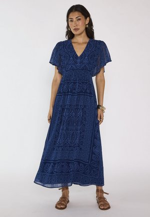 Femme debout, portant une longue robe bleue fluide à motifs avec des manches volantées, sandales brunes, bijoux en or, fond neutre.