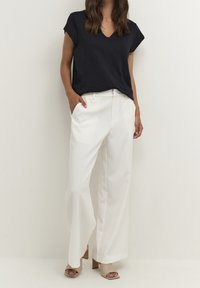 Marinblå v-ringad t-shirt kombinerad med vita vida byxor. Byxorna har en mjuk textur och modellen bär beige klacksandaler.