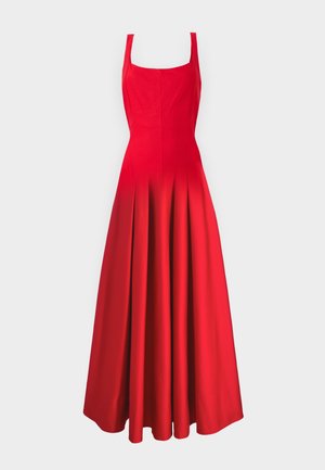 Femme debout portant une robe longue rouge sans manches avec un décolleté carré et des sandales à talons hauts noires à brides, sur un fond blanc.
