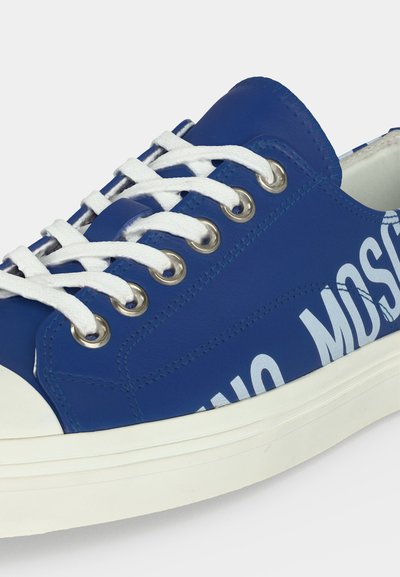 Baskets basses bleues avec lacets blancs, œillets métalliques, semelle en caoutchouc blanche et lettrage blanc partiel sur le côté.