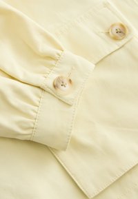 Gros plan sur un tissu jaune clair avec deux boutons beiges sur un poignet de manche froncé et un rabat de poche.
