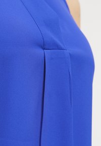 Kuninkaansininen paita, jossa on sileä, mattapintainen kangas; sivussa laskos lisää yksityiskohtia. Lähikuva korostaa rakennetta ja väriä.