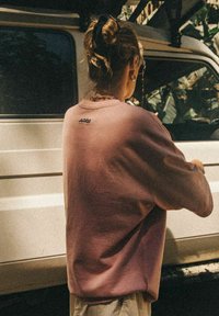 Jasnoróżowy, oversize'owy sweatshirt z okrągłym dekoltem, ściągaczami na rękawach i małym logo na plecach. Materiał wydaje się miękki i teksturowany.