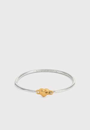 Dünnes silbernes Armband mit einem kleinen goldenen herzförmigen Verschluss in der Mitte auf einem schlichten weißen Hintergrund.