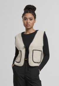 Béžová fleecová vesta s černými lemy, s véčkovým výstřihem, dvěma předními kapsami a zavazováním vpředu.