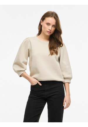 Femme aux longs cheveux bruns posant avec une main dans la poche, portant un haut beige à manches bouffantes et un pantalon noir sur fond blanc.