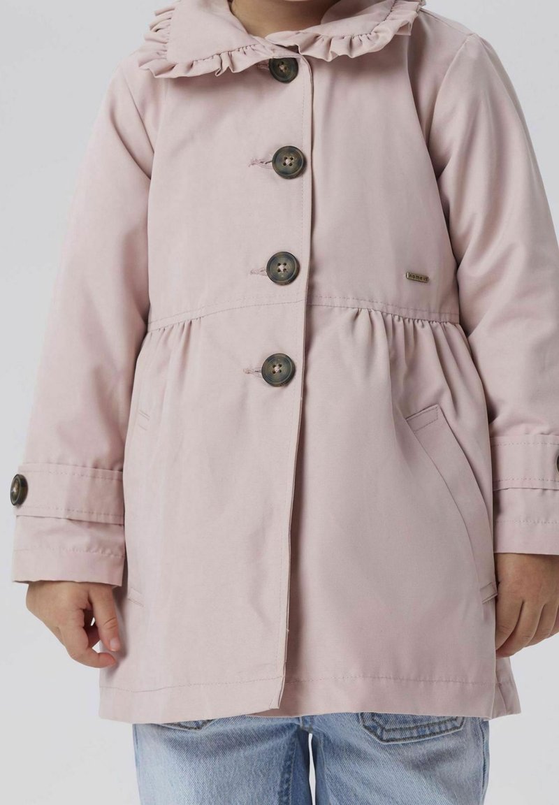 Enfant portant un manteau rose clair avec des boutons foncés et un col à volants, debout les mains le long du corps, portant un jean bleu clair.