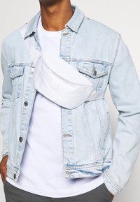 Vit tygmidjeväska med en böjd design och en framficka med dragkedja, bärs över en ljusblå jeansjacka, med justerbara axelremmar.