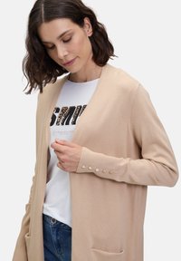 Beige gebreide cardigan met een open voorkant, geribbelde manchetten met knoopaccenten en voorzakken, gedragen over een witte graphic t-shirt.