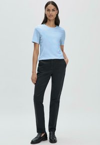 Vaaleansininen puuvillapaita lyhyillä hihoilla, yhdistettynä mustiin muotonsa mukaan valmistettuihin housuihin. Malli seisoo toisessa kädessä taskussa, jalassaan mustat loafertossut.