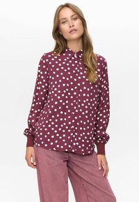Femme portant une blouse boutonnée à manches longues maroon avec des pois blancs et un pantalon large rose devant un fond blanc.