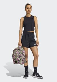 Top corto nero e pantaloncini, abbinati a scarpe da ginnastica nere. Uno zaino con motivo floreale con accenti verdi e viola è tenuto in una mano.