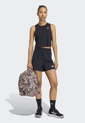 Schwarzes bauchfreies Oberteil und Shorts, kombiniert mit schwarzen Sneakern. Ein Rucksack mit floralem Muster und grünen sowie lila Akzenten wird in einer Hand gehalten.