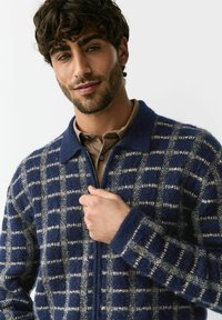 Námornícka modrá a krémová károvaná pletená bunda s golierom, zapínaním na zips a textúrovanou látkou so zvýraznenými obdĺžnikovými vzormi.