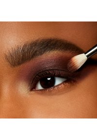 Gros plan sur un œil avec un fard à paupières bourgogne riche, complété par un doux éclaircissement doré. Un pinceau estompé applique le maquillage le long de la paupière.