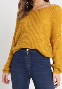 Yellow knit sweater with an off-the-shoulder design, textured ribbing, and loose fit, paired with high-waisted dark blue jeans featuring a zipper.