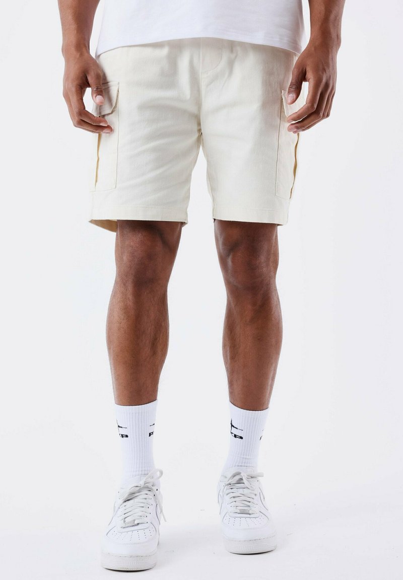 Pantalones cortos de carga para hombre en color crema con bolsillos laterales, detalles en amarillo contrastante, corte ajustado, material de algodón, combinados con calcetines blancos y zapatillas.}}