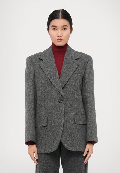 Blazer en écossais gris avec un bouton unique, coupe ajustée, larges revers et deux poches avant, porté par-dessus un col roulé bordeaux.