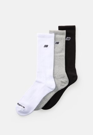 Trois paires de chaussettes de sport New Balance en blanc, gris et noir, chacune arborant le logo de la marque près du haut du revers, disposées en chevauchement.