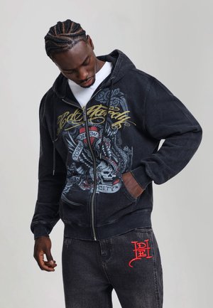 Hombre con el pelo trenzado que lleva una sudadera negra con cremallera con diseño de calavera y alas y vaqueros negros con logotipo bordado en rojo.