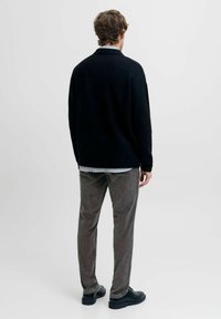 Suéter negro de punto, pantalones de pana gris, zapatos negros, dobladillo de la camisa visible. Modelo visto desde atrás contra un fondo claro y liso.