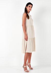 Knee-length cream dress with a textured finish, featuring a fitted bodice and a flared skirt. Paired with beige high-heeled sandals.